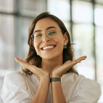 Cheerful business woman with glasses posing with her hands under her face showing her smile in an office. Playful hispanic female entrepreneur looking happy and excited at workplace